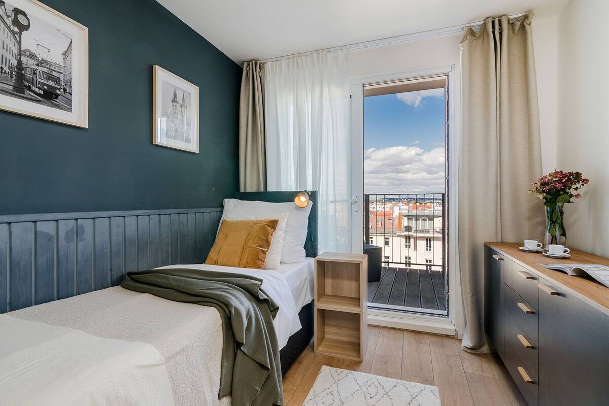 Ein Schlafzimmer mit Bett, Nachttisch, Kommode, einem Fenster mit Vorhängen und einem Balkon mit Blick auf Gebäude.