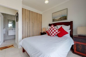 A bedroom with a wooden headboard, a large bed with white bedding, red and white checkered pillows, a nightstand with a lamp, and a painting on the wall.