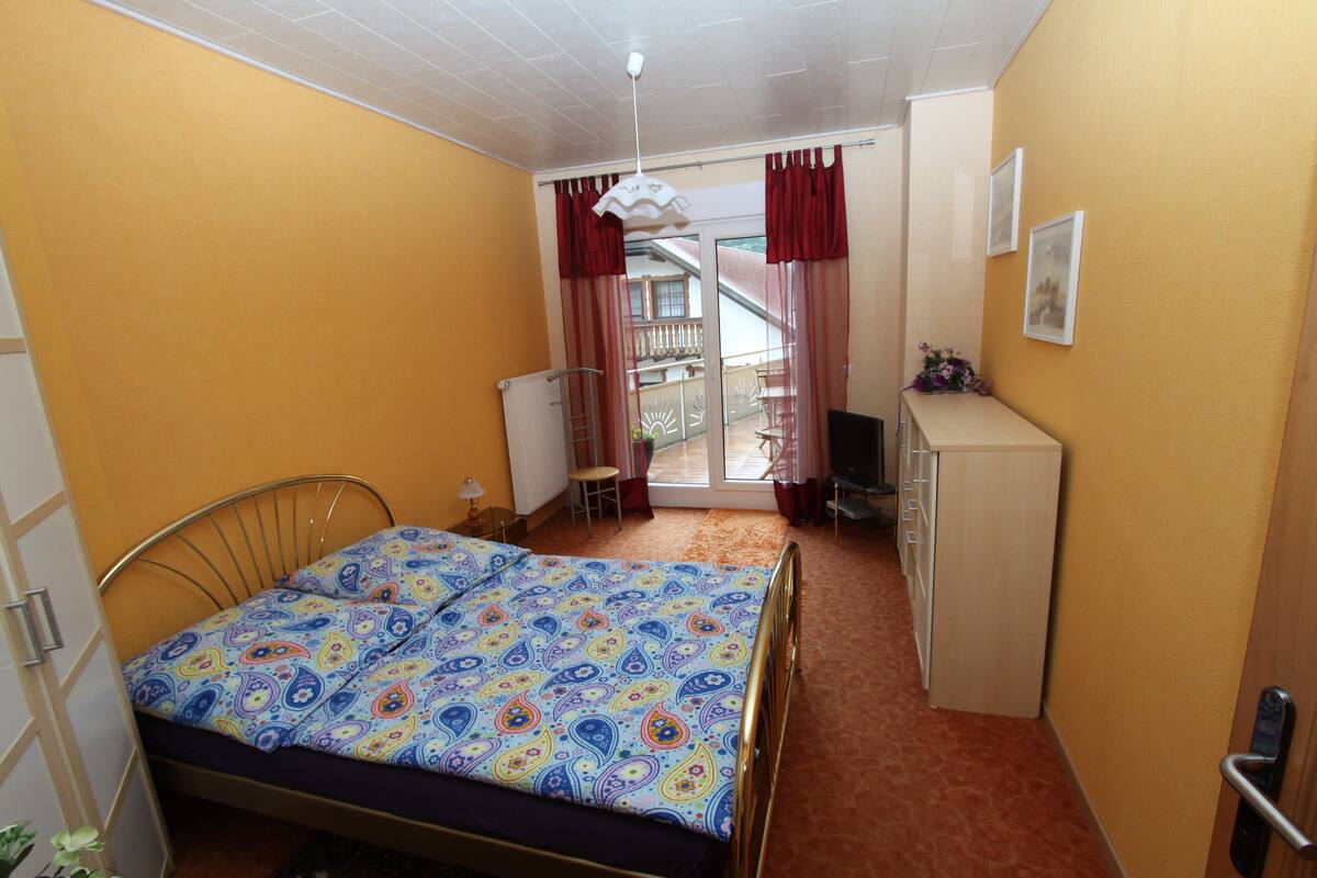 A small, brightly lit room with a bed, a desk, and a chair.
