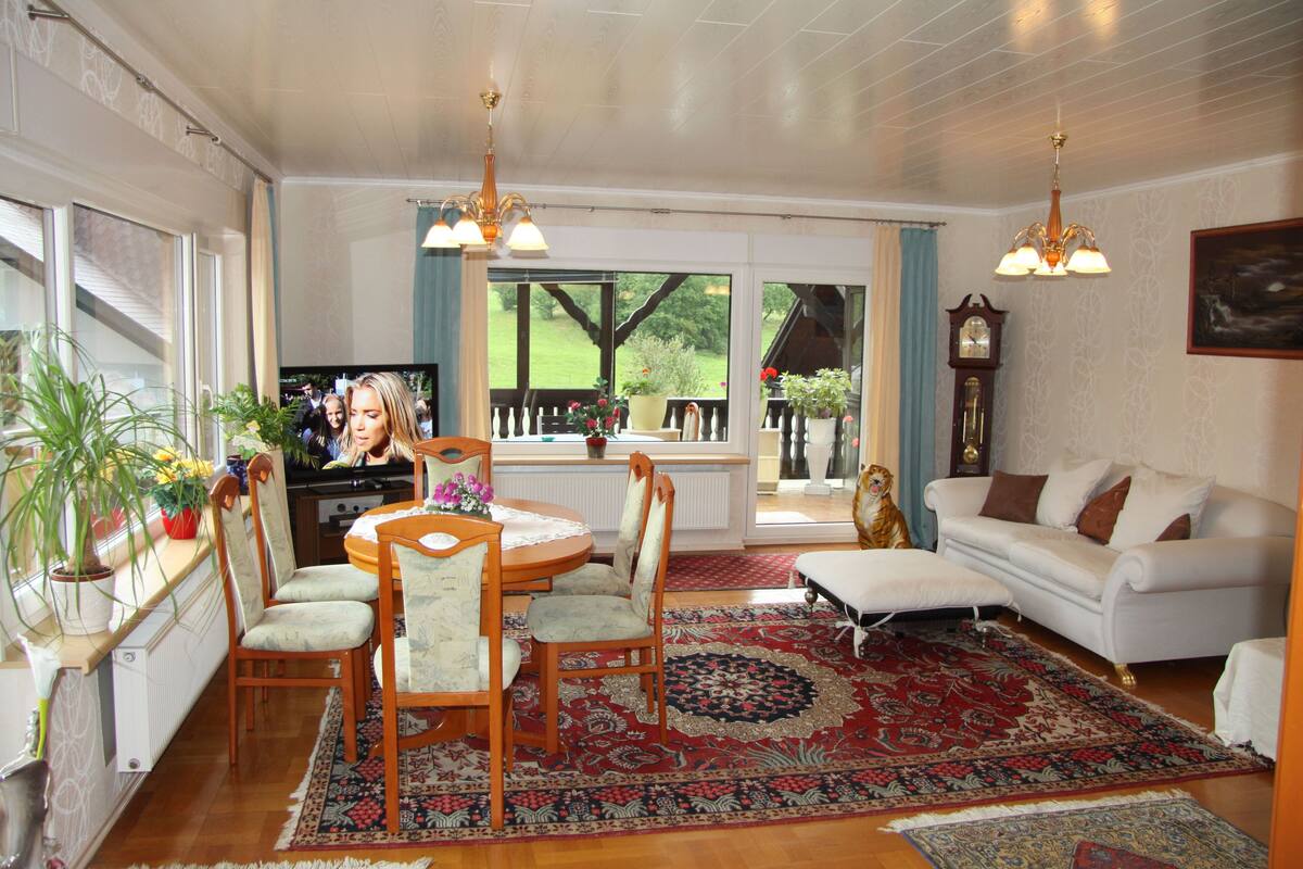 A living room with a dining table, chairs, a sofa, a TV, and a large window with a view of greenery.