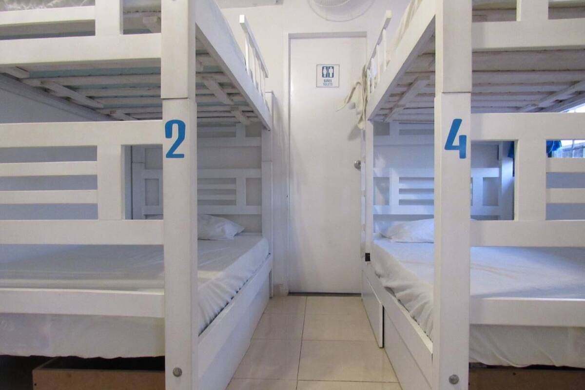 Un couloir étroit avec des lits superposés numérotés 2 et 4, une porte portant un panneau indiquant la sortie de secours et un sol carrelé.