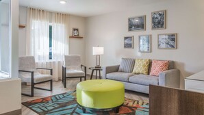 A modern living room with a sofa, two armchairs, a green ottoman, a patterned rug, and a window with sheer curtains. - WoodSpring Suites Atlanta McDonough (McDonough)