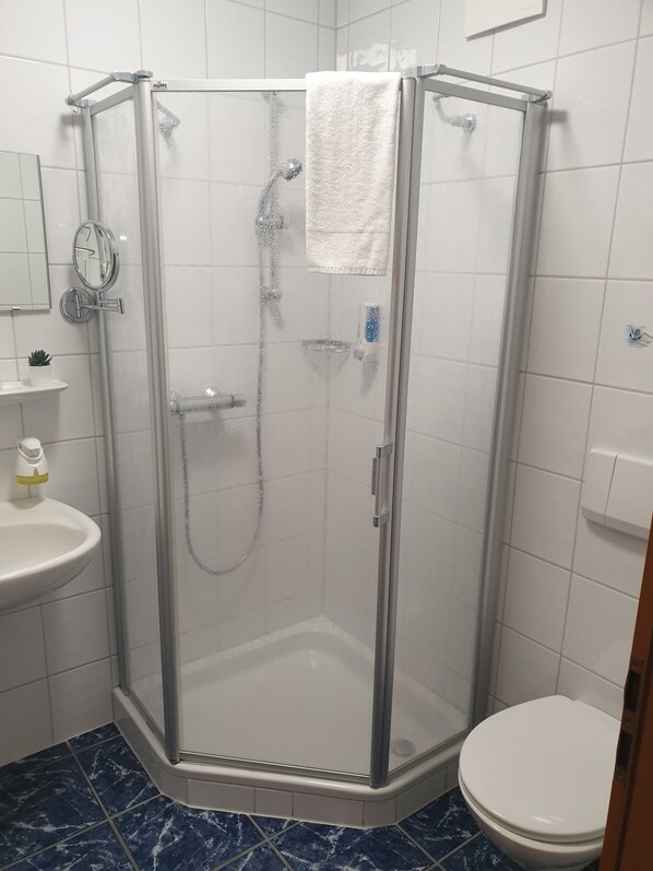 A glass-enclosed shower stall with a white towel hanging on the door handle, a handheld showerhead, and a handheld faucet. - Blischkes Hotel (Wittmund)