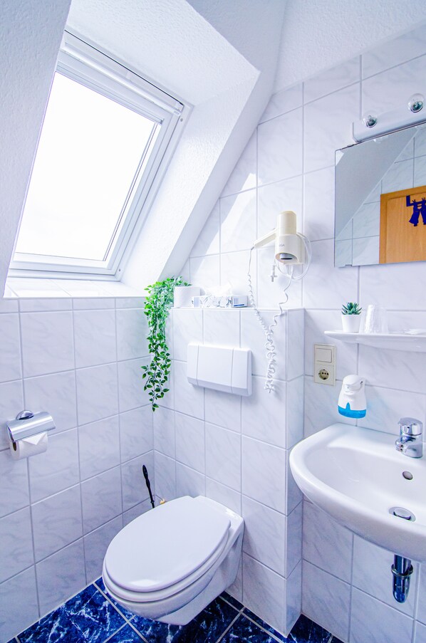 A bathroom with a skylight, white tiles, a toilet, a sink, and a mirror. - Blischkes Hotel (Wittmund)