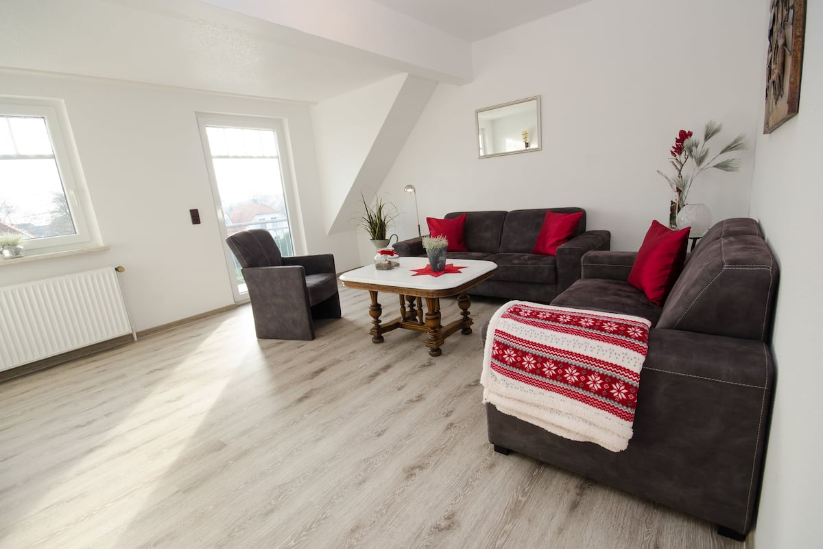 A modern living room with a brown sofa, a round coffee table, and a grey armchair. There is a window on the left and a mirror on the wall.