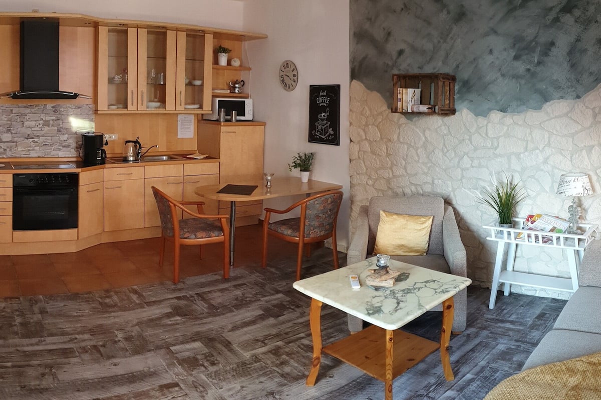 A compact kitchen with wooden cabinets, a microwave, and a stove. There is a dining area with a table and chairs. The room features a stone wall and a staircase.