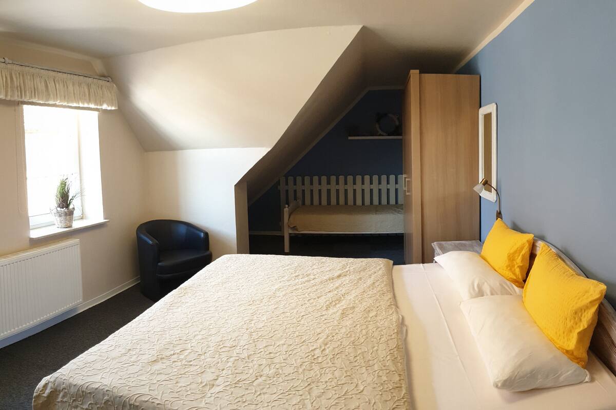 A bedroom with a bed, a chair, a window, a staircase, and a wall-mounted lamp.