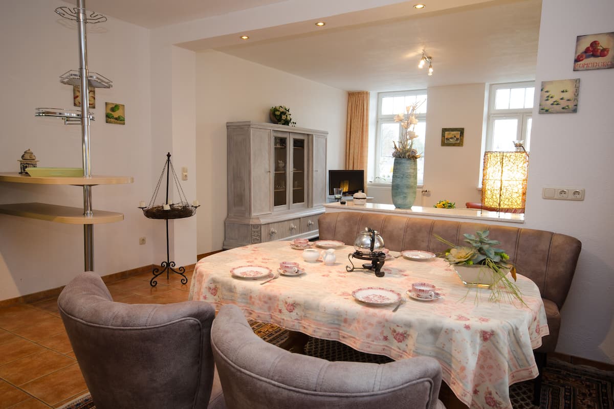 A dining area with a round table covered in a floral tablecloth, surrounded by cushioned chairs. A cabinet with glass doors and a hanging light fixture are visible in the background.