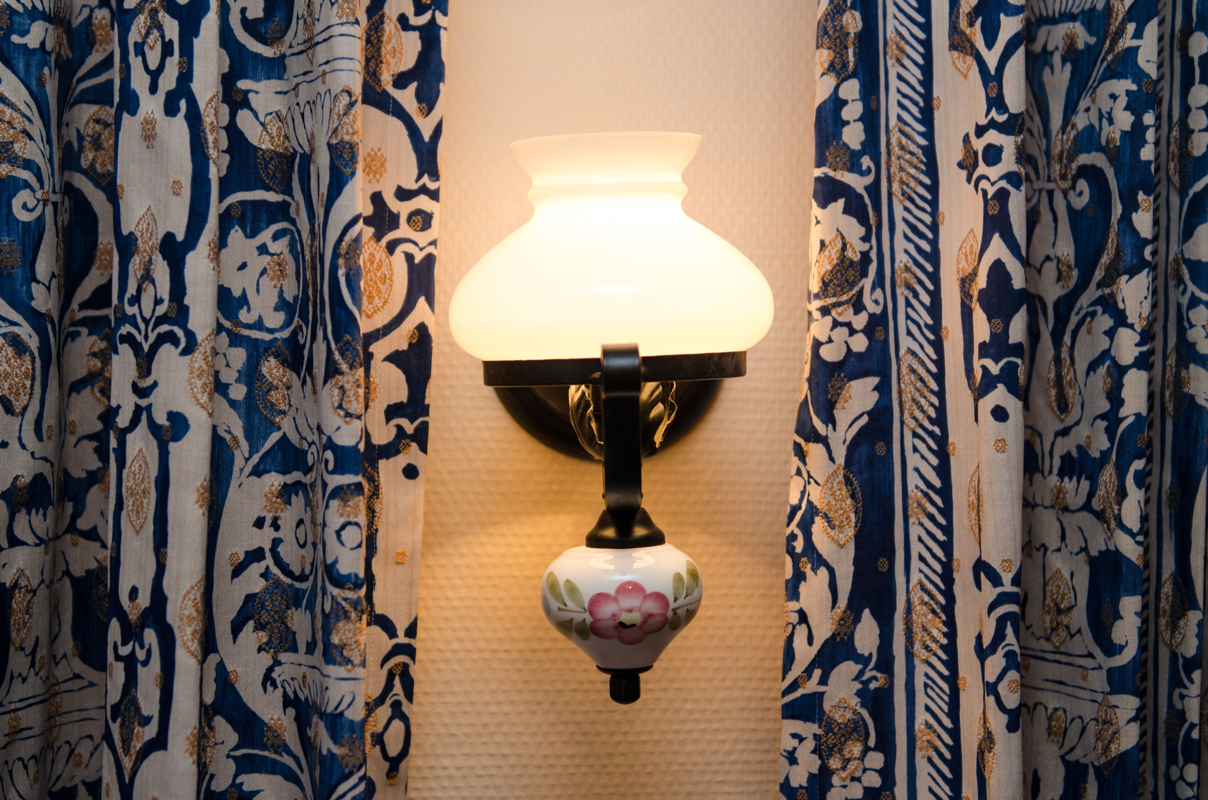 Una lámpara de pared con diseño floral, ubicada entre cortinas estampadas en azul y blanco.