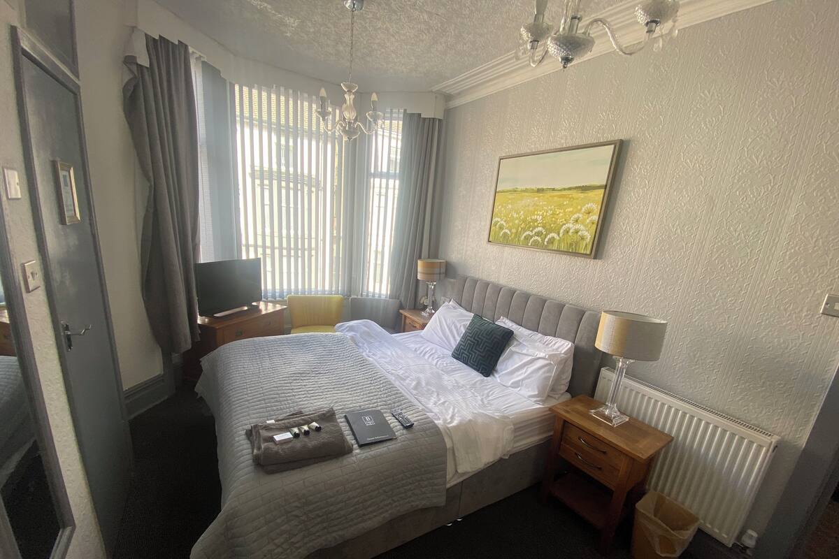 Ein Hotelzimmer mit einem großen Bett, einem Nachttisch, Vorhängen an einem Fenster, einer Chandelieber und einem Gemälde an der Wand.