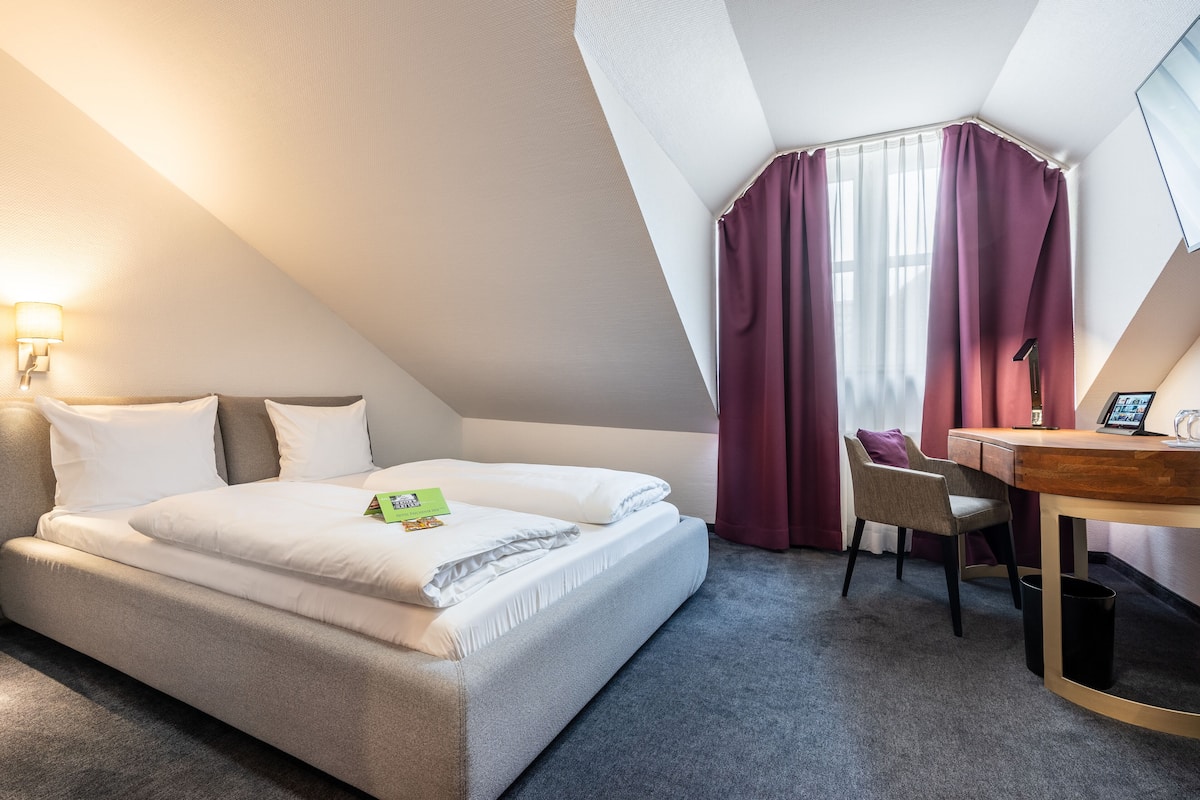 A hotel room with a bed, a desk, a chair, and a window with curtains.