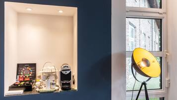 A hotel room with a coffee station, a wall-mounted shelf with cups and a coffee machine, and a window with a view of buildings.