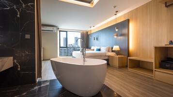 A modern hotel room with a large bathtub, a bed with blue pillows, a wooden wall, and a view of the city.