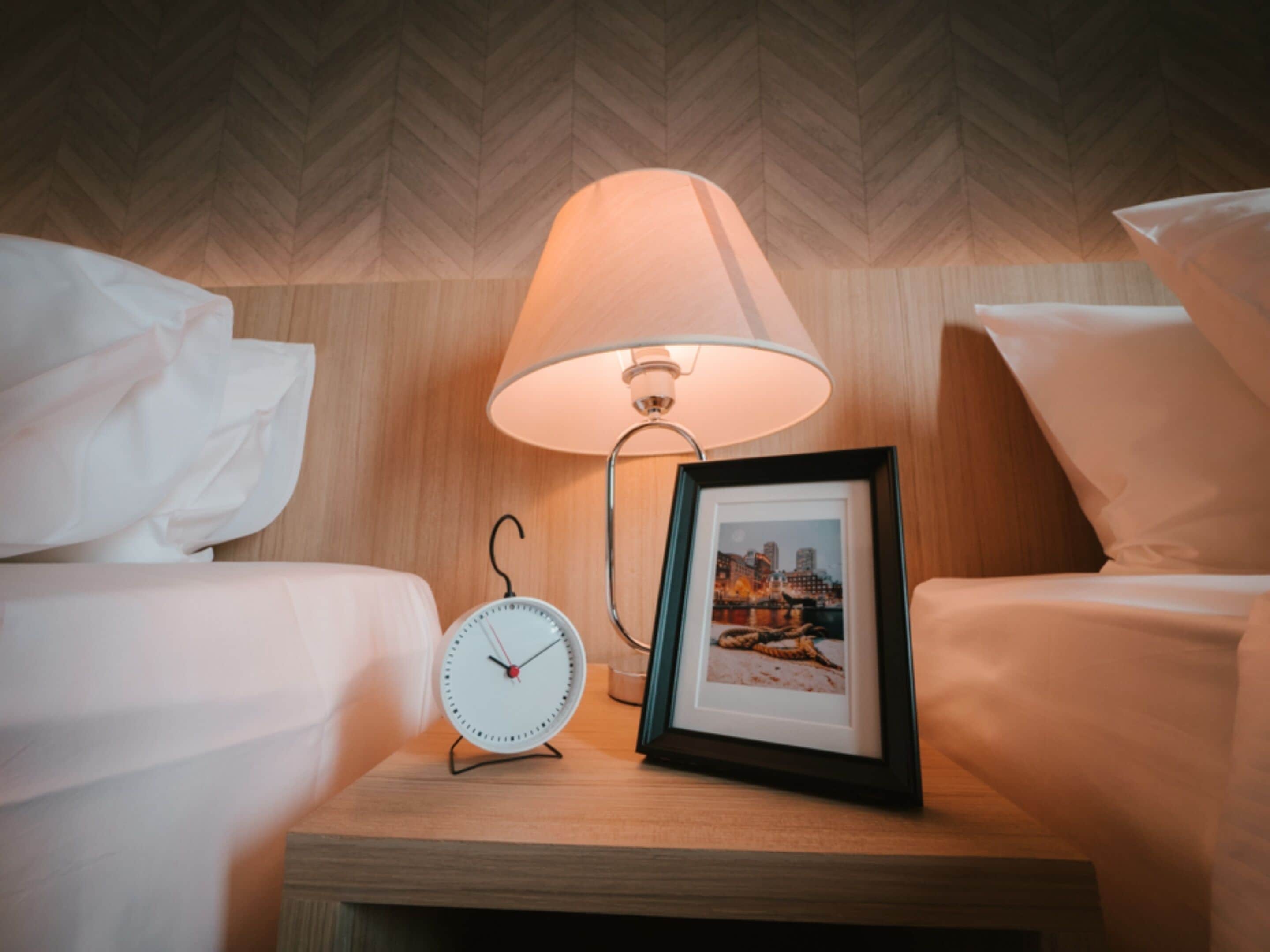 Een net opgemaakt bed met witte lakens, een nachtkastje met een ingelijst foto, een klok en een lamp.