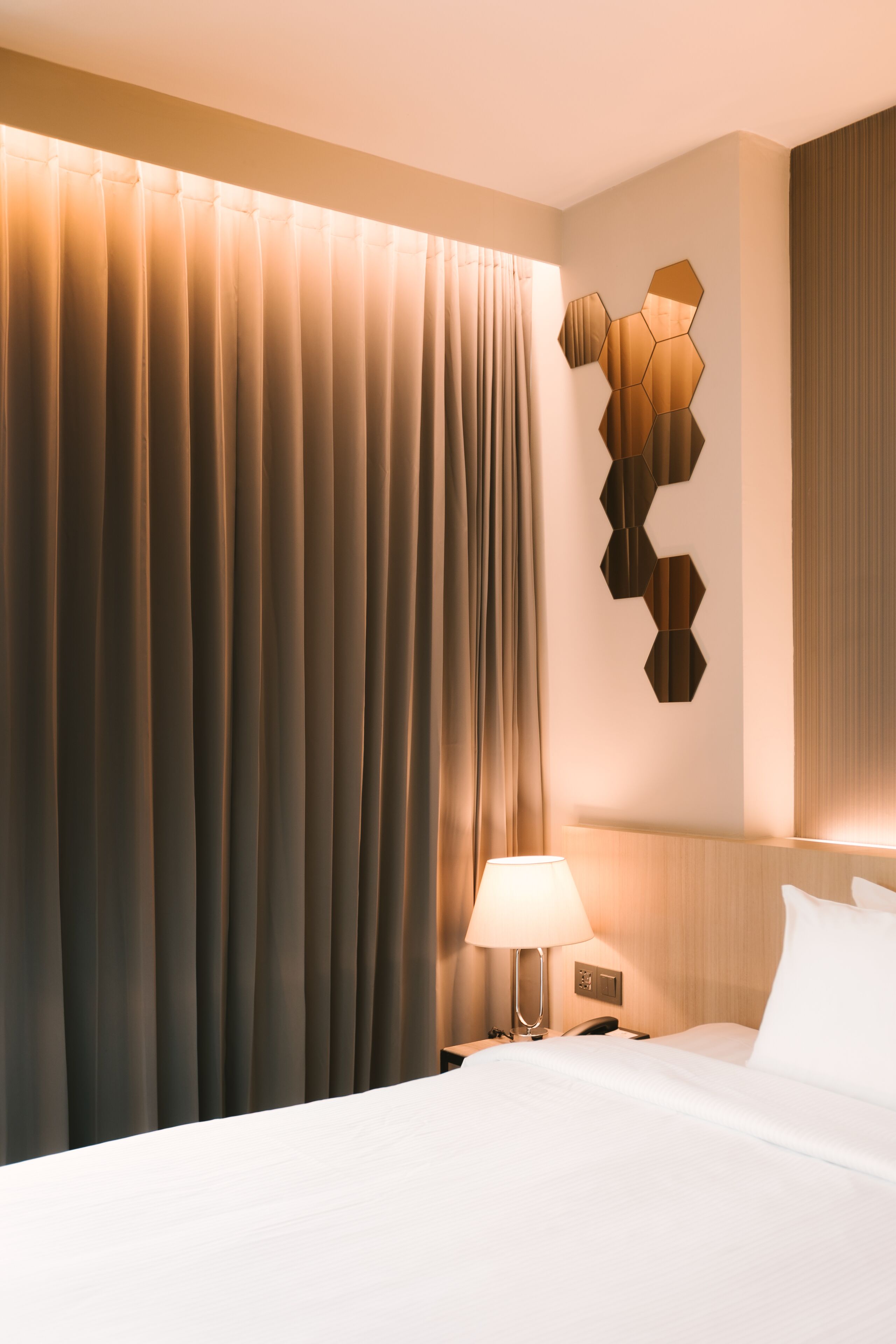 Una habitación de hotel con una cama grande, una lámpara de noche, un adorno geométrico en la pared y una cortina.