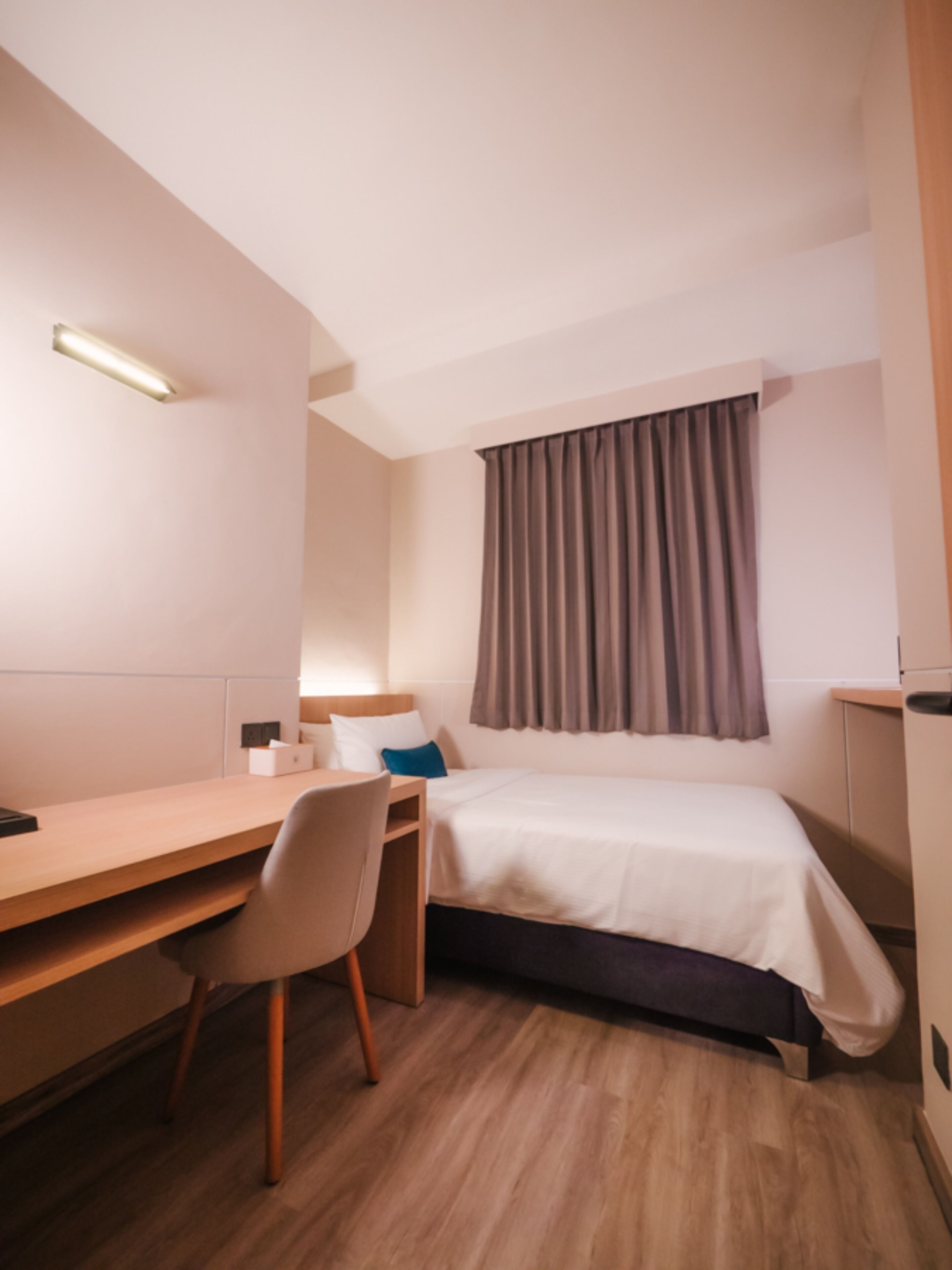 A hotel room with a bed, a desk, a chair, a bedside table, and a window with curtains.