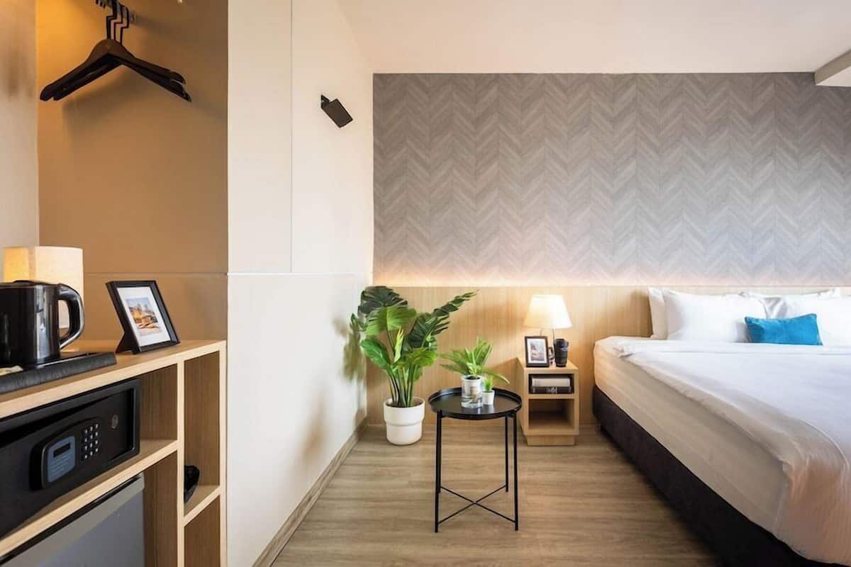 A hotel room with a bed, bedside table, lamp, potted plants, and a desk with a coffee maker.