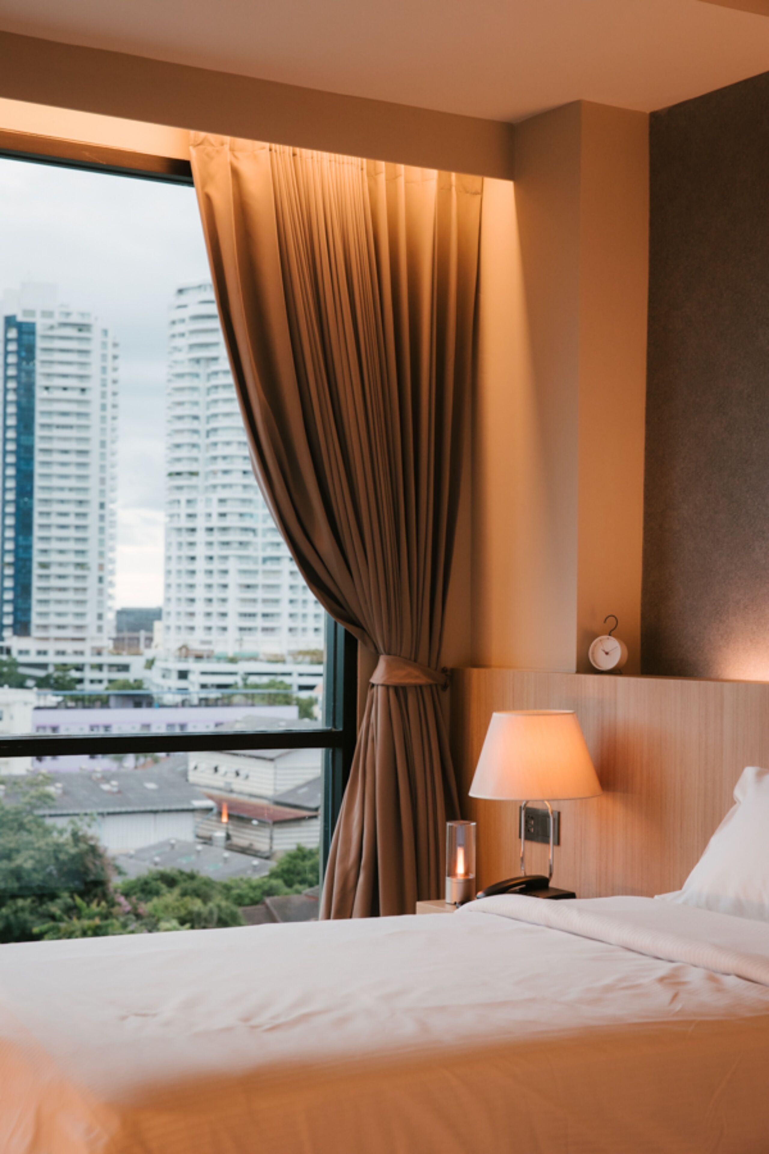 A hotel room with a bed, a nightstand with a lamp, a clock, and a view of tall buildings through a window with curtains.