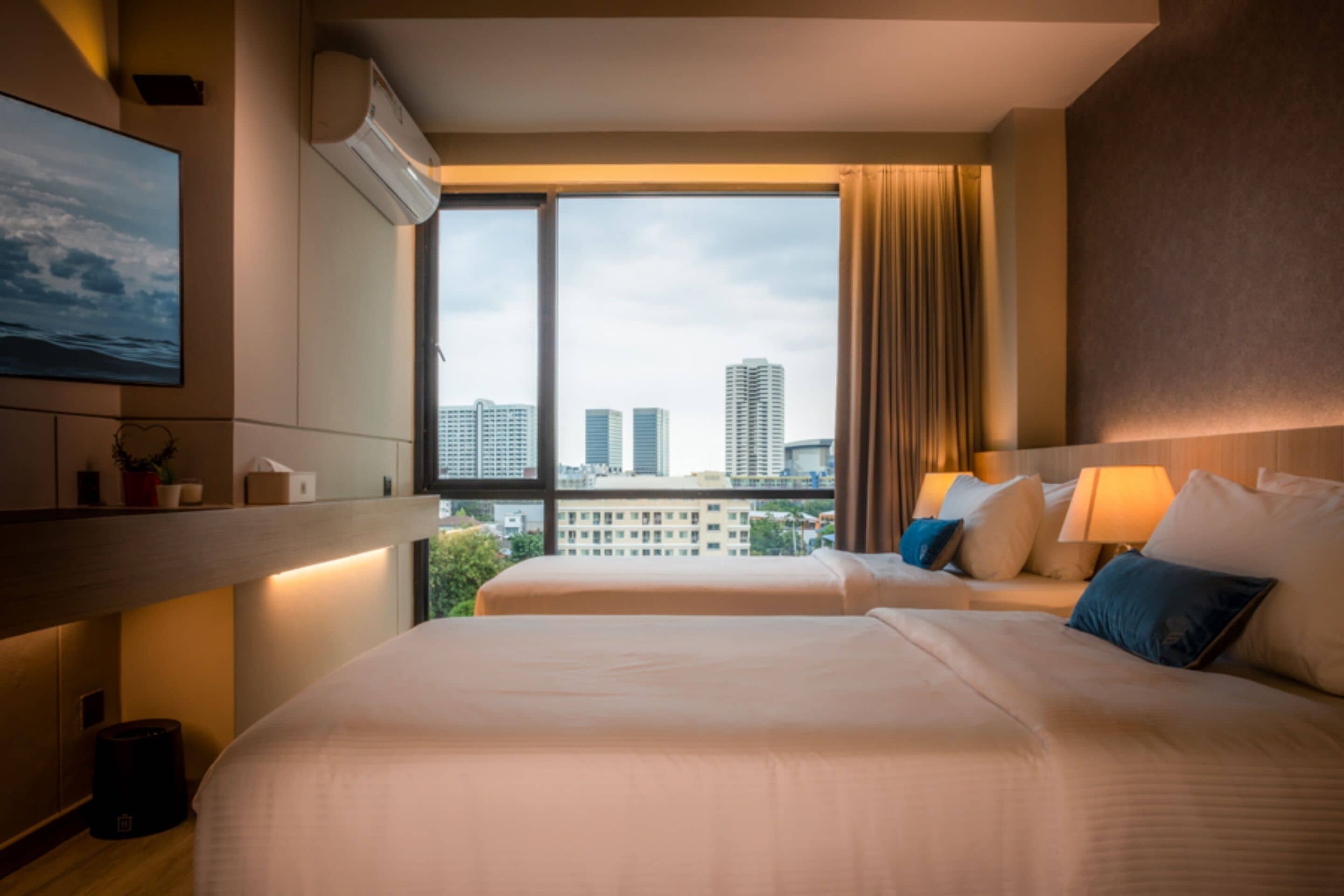 Habitación de hotel con dos camas, un ventanal con vistas a la ciudad, un equipo de aire acondicionado y una mesita de noche con lámpara.