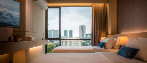 Habitación de hotel con dos camas, un ventanal con vistas a la ciudad, un equipo de aire acondicionado y una mesita de noche con lámpara.