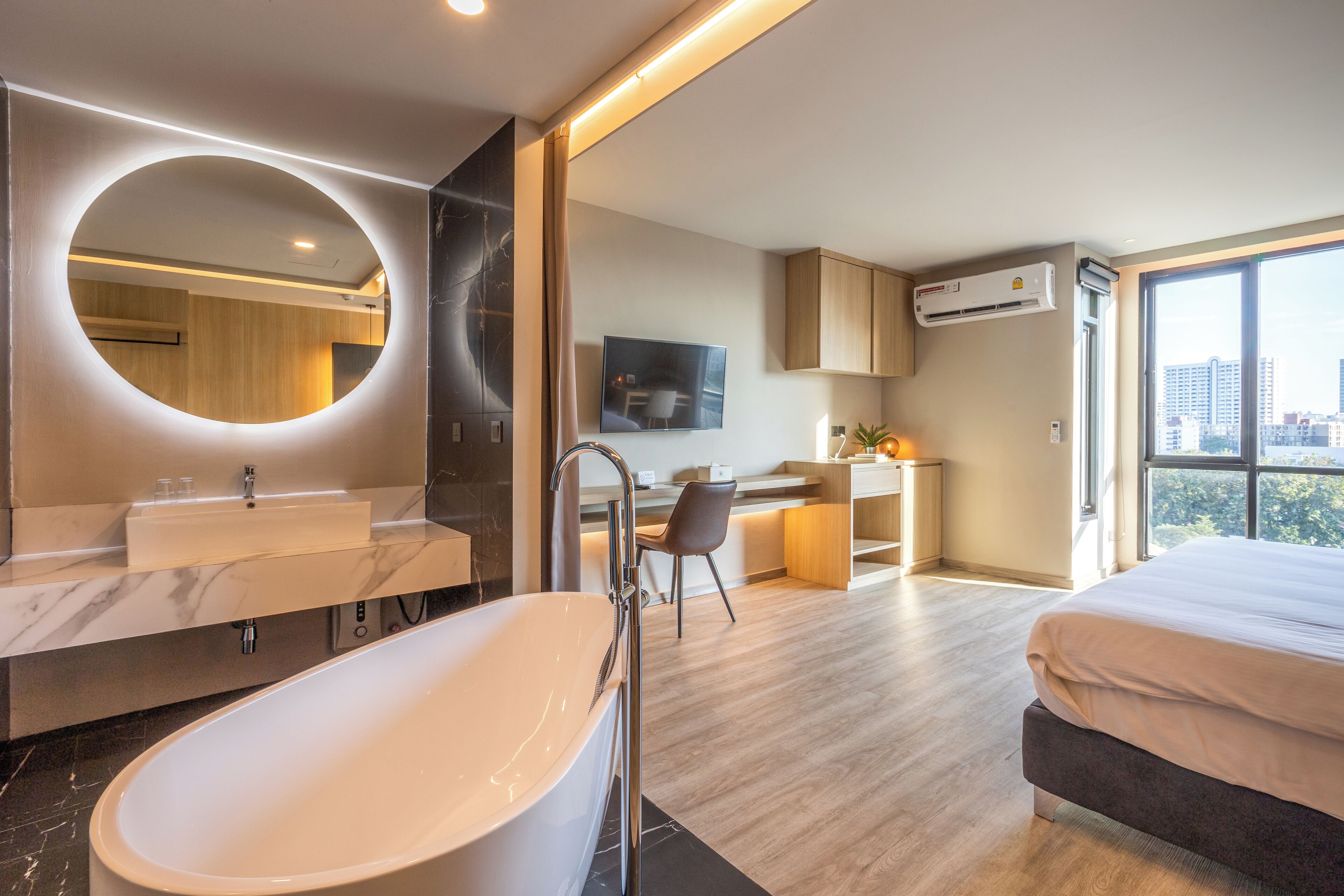 A modern hotel room with a large oval bathtub, a round mirror, a desk with a chair, and a large window offering a city view.