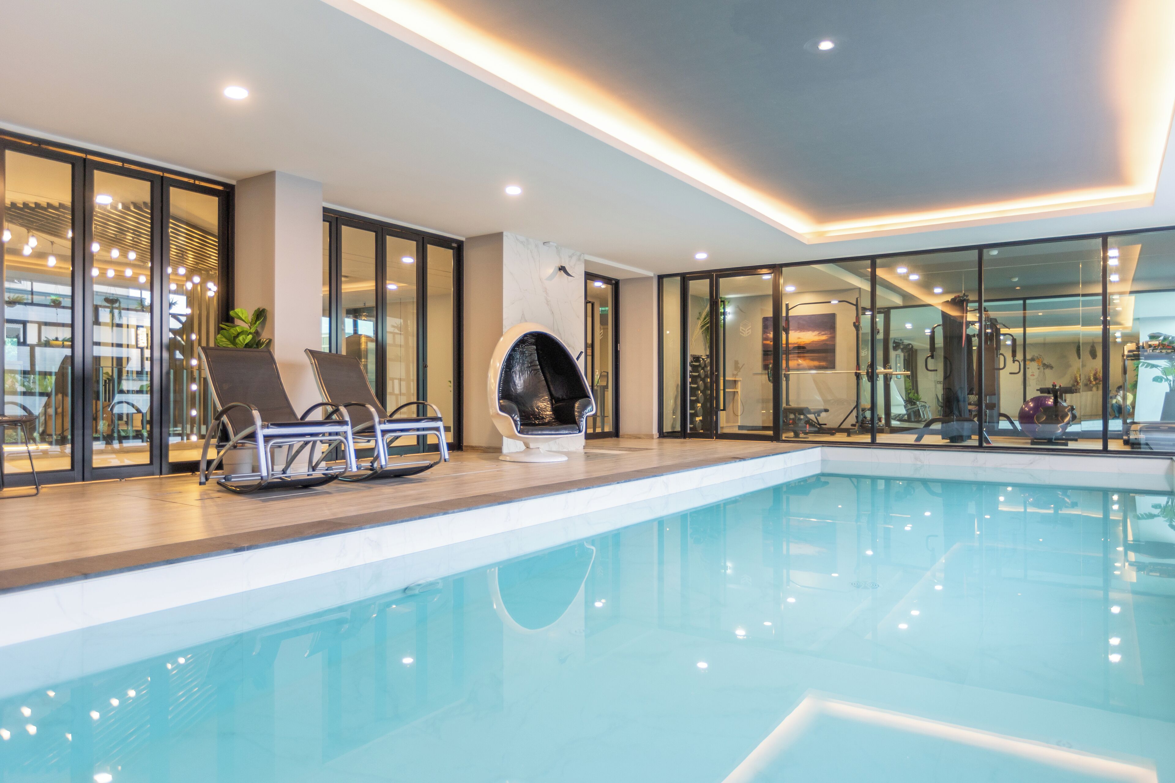 A modern indoor pool area with glass walls, lounge chairs, and a ceiling with recessed lighting.