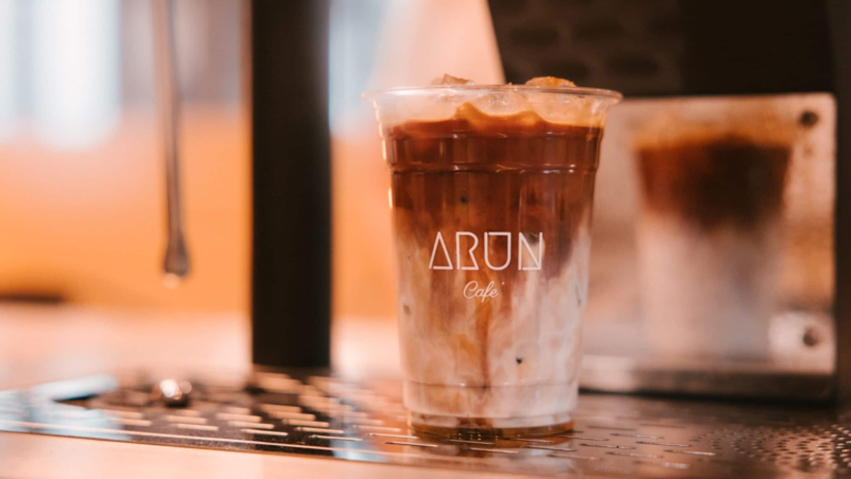 A clear glass of iced coffee with ice cubes, labeled "ARUN Cafe," on a metal surface with a blurred background.