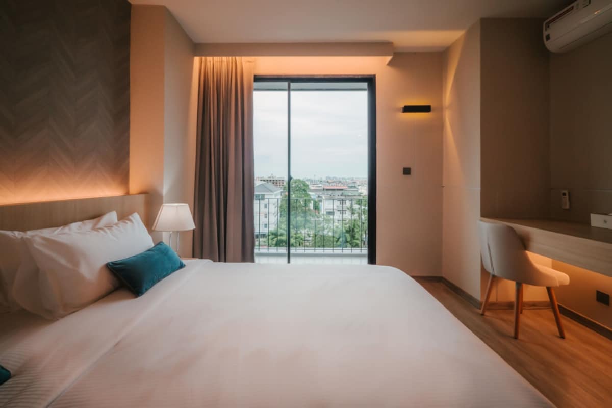 A hotel room with a bed, a desk, a chair, a window with a view of buildings, and an air conditioning unit.