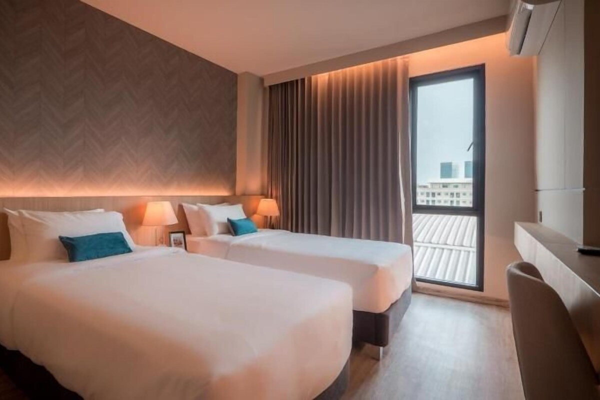 A hotel room with two beds, a large window with curtains, a desk, and a view of buildings.