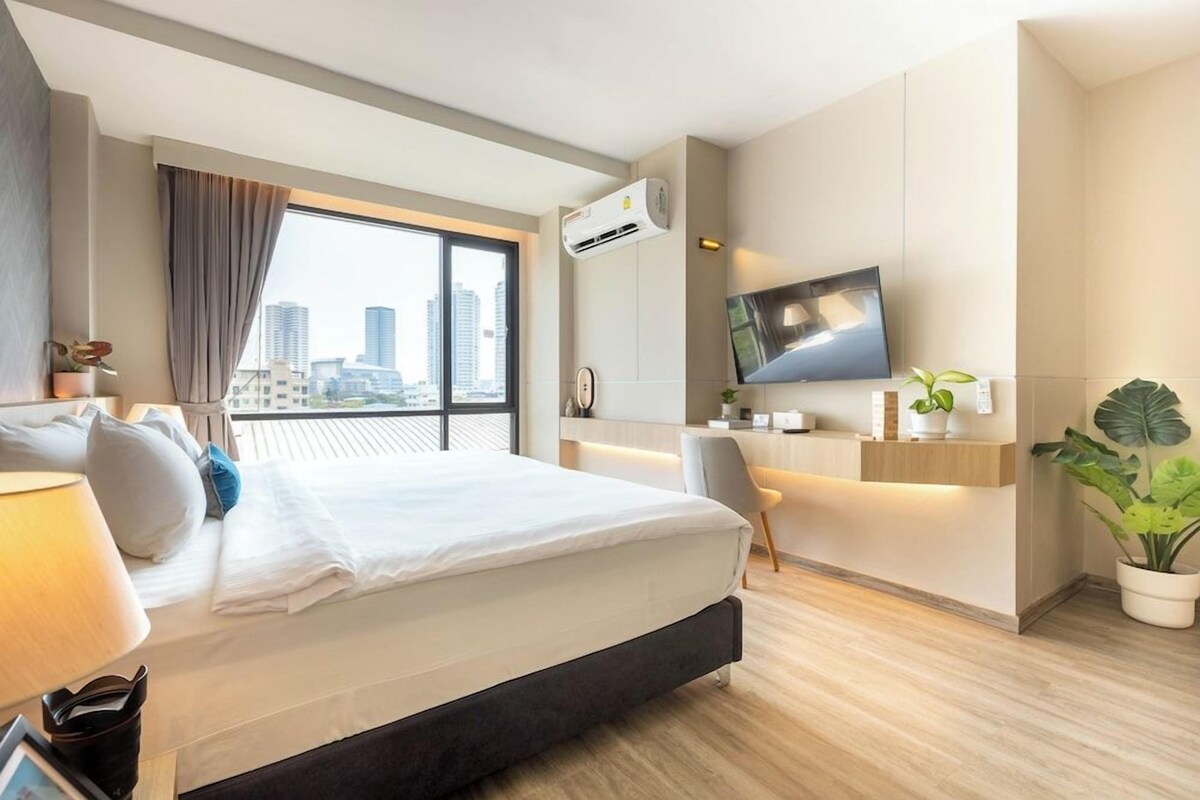 A modern hotel room with a large bed, a desk, a flat-screen TV, and a city view.