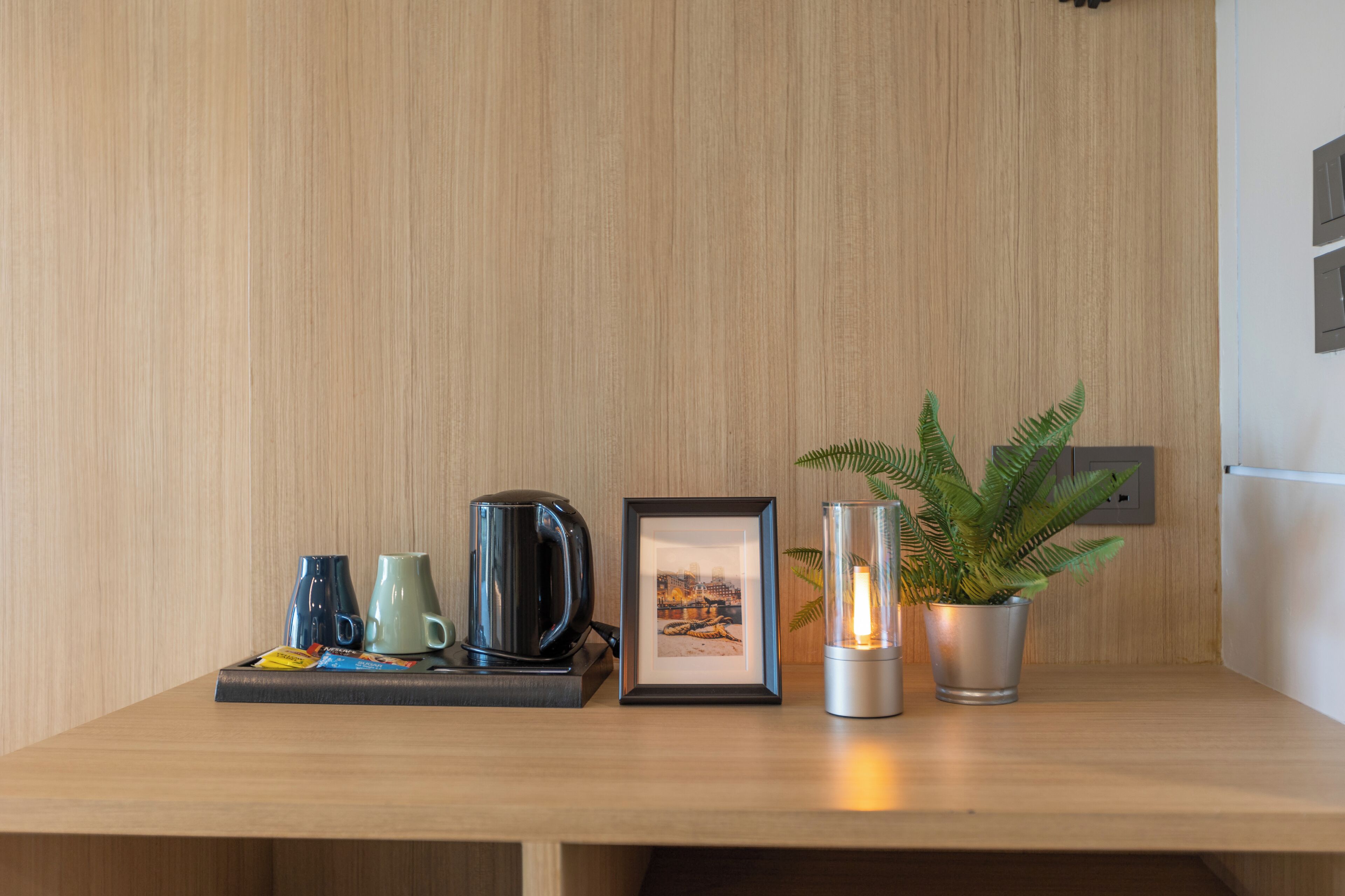A wooden table with a framed picture, a potted plant, a candle, and a tray with cups and a kettle.