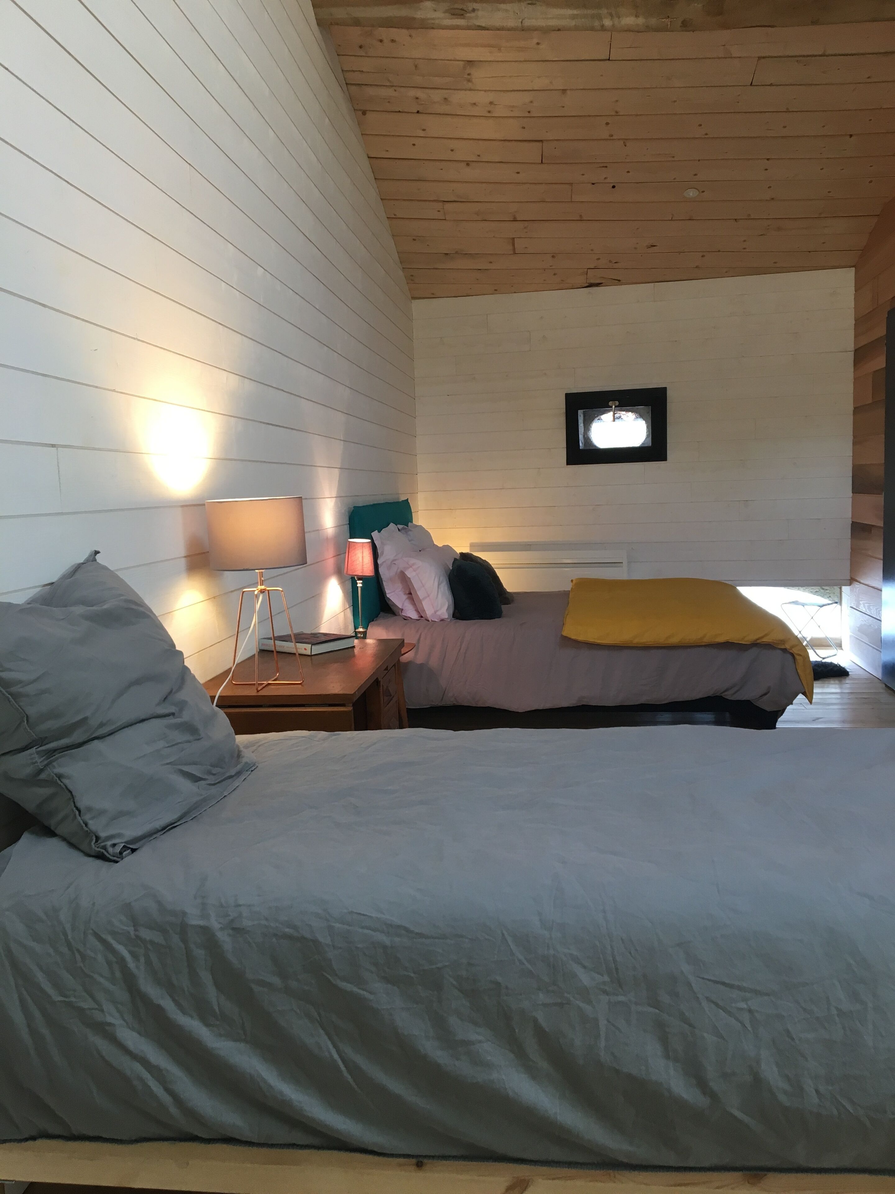 A bedroom with a wooden ceiling, a bed with a gray blanket, a bedside table with a lamp, and a framed picture on the wall.