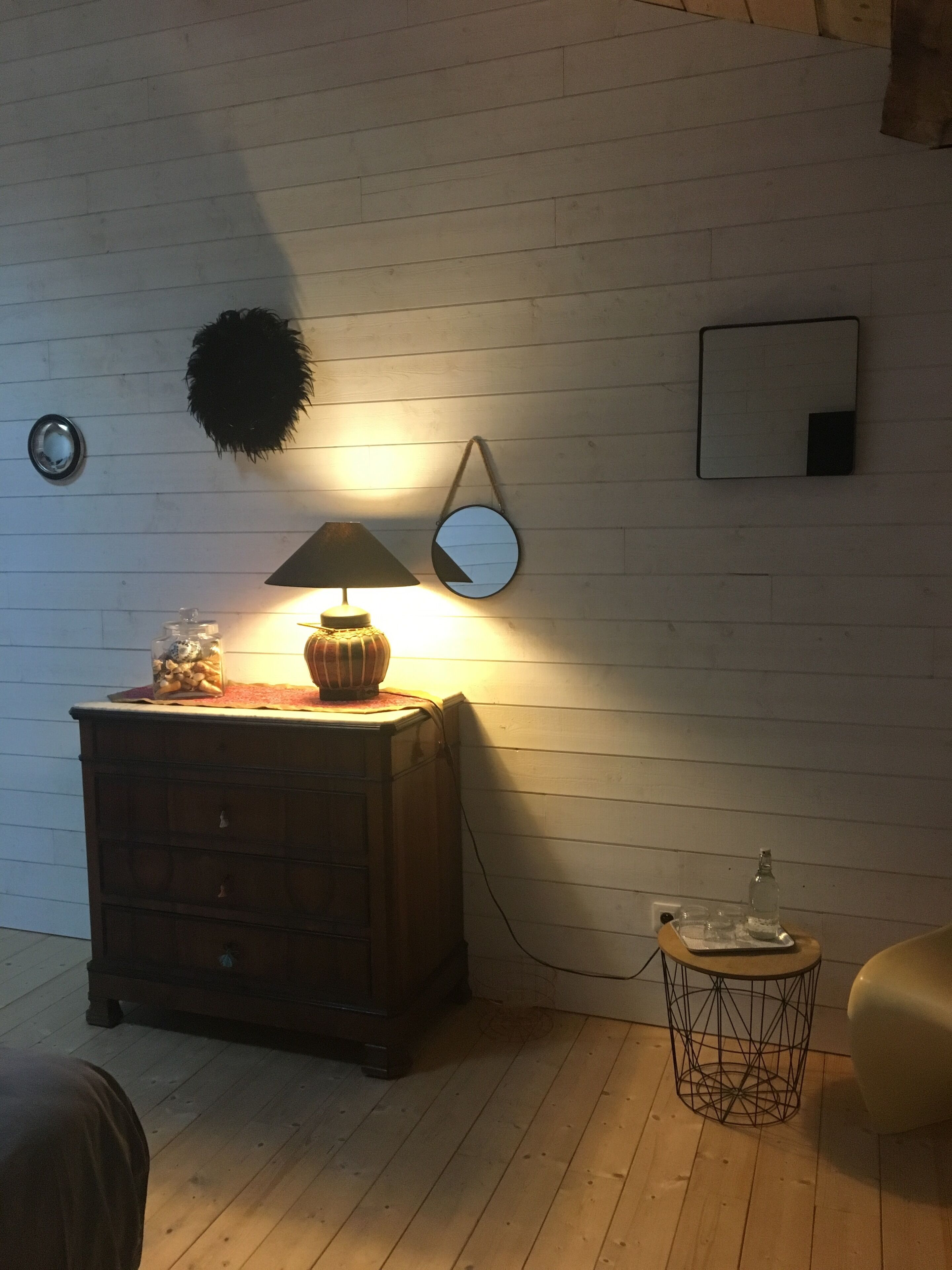 Et værelse med et træmøbel, en lampe, et rundt spejl og et lille rundt bord med en flaske og glas.