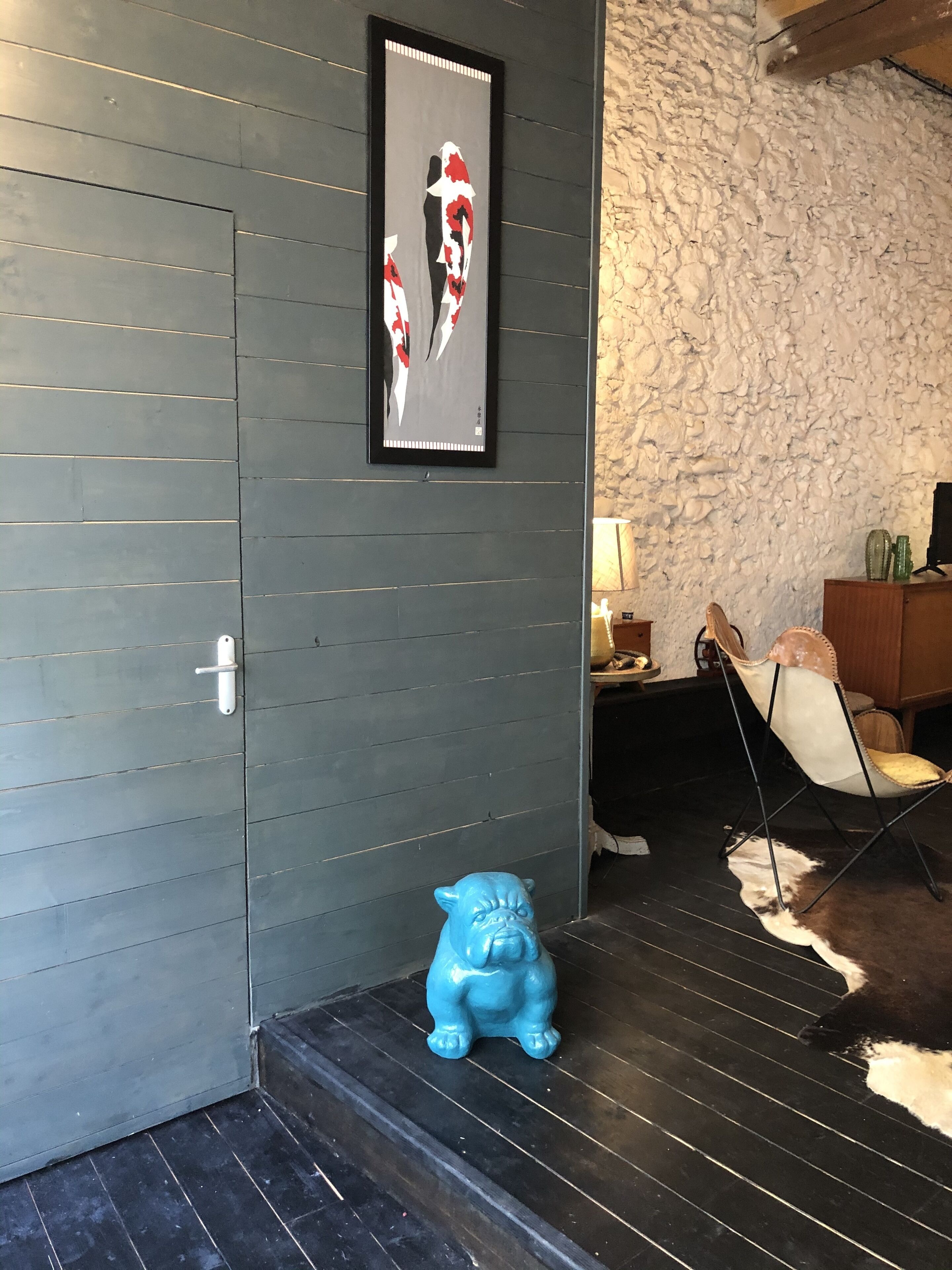 A room with a grey wooden wall, a framed artwork, a blue ceramic pig, a chair, and a cowhide rug.