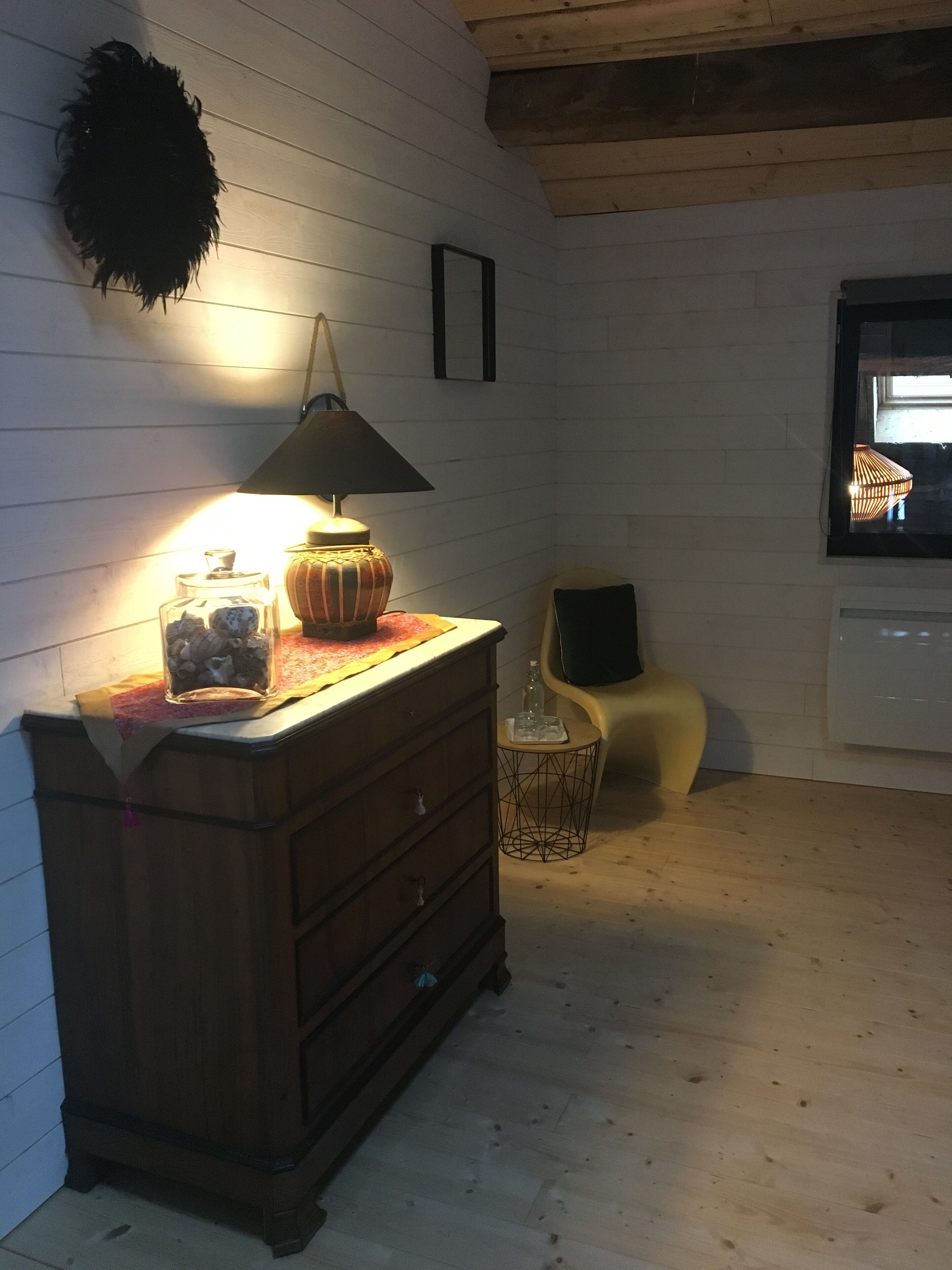 Quarto com cômoda de madeira, luminária, mesa pequena com uma garrafa, cadeira e janela com vista.