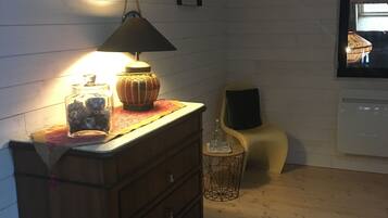 Quarto com cômoda de madeira, luminária, mesa pequena com uma garrafa, cadeira e janela com vista.