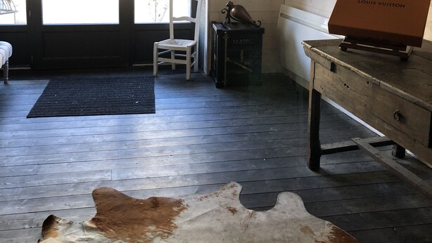 Una habitación con piso de madera, un tapete de cuero de vaca, una silla blanca, un escritorio con una caja Louis Vuitton y una pared con adornos.