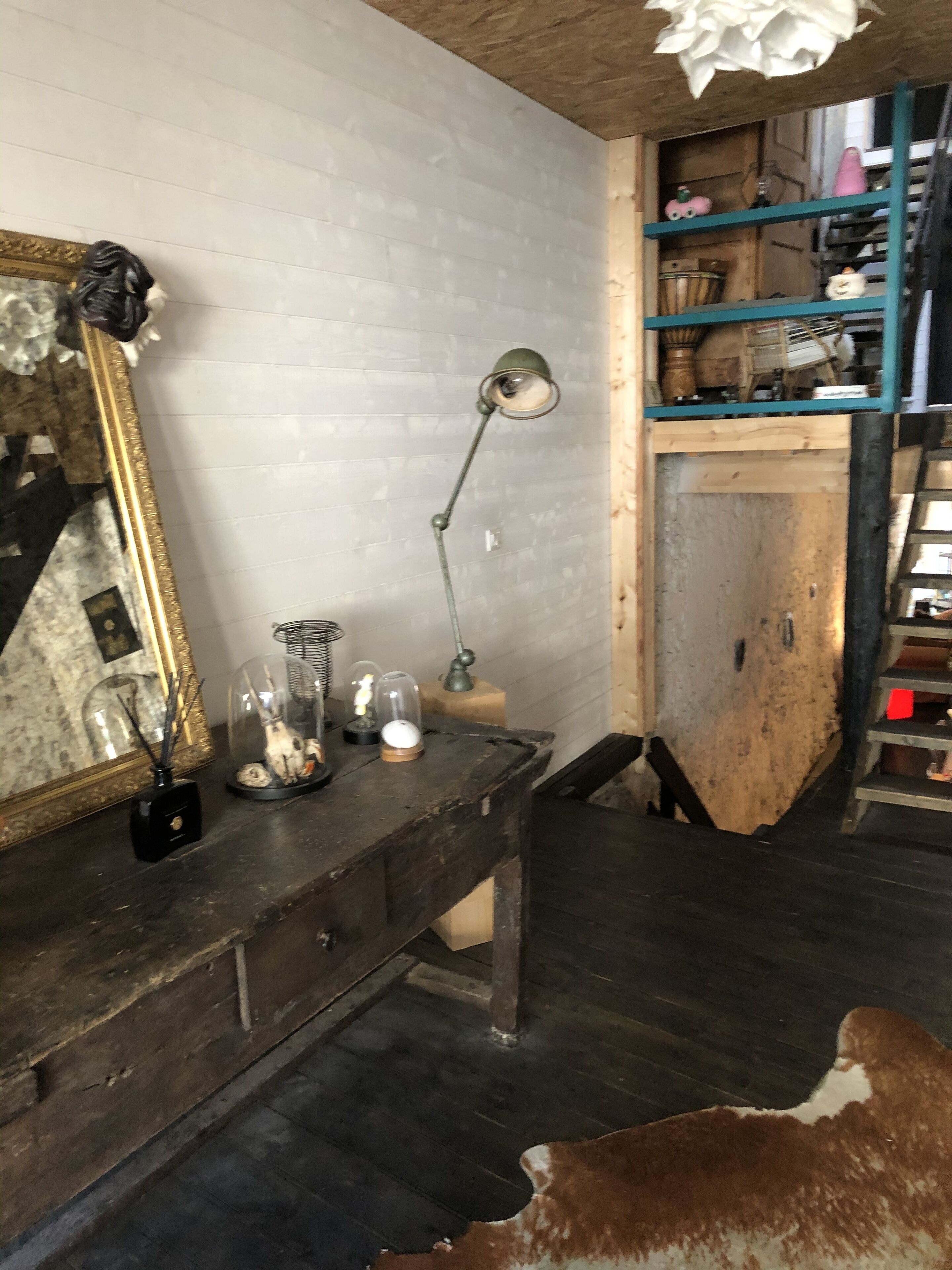 Una habitación rústica con un escritorio de madera, un espejo, una lámpara, estantes con diversos objetos y una alfombra de cuero de vaca.