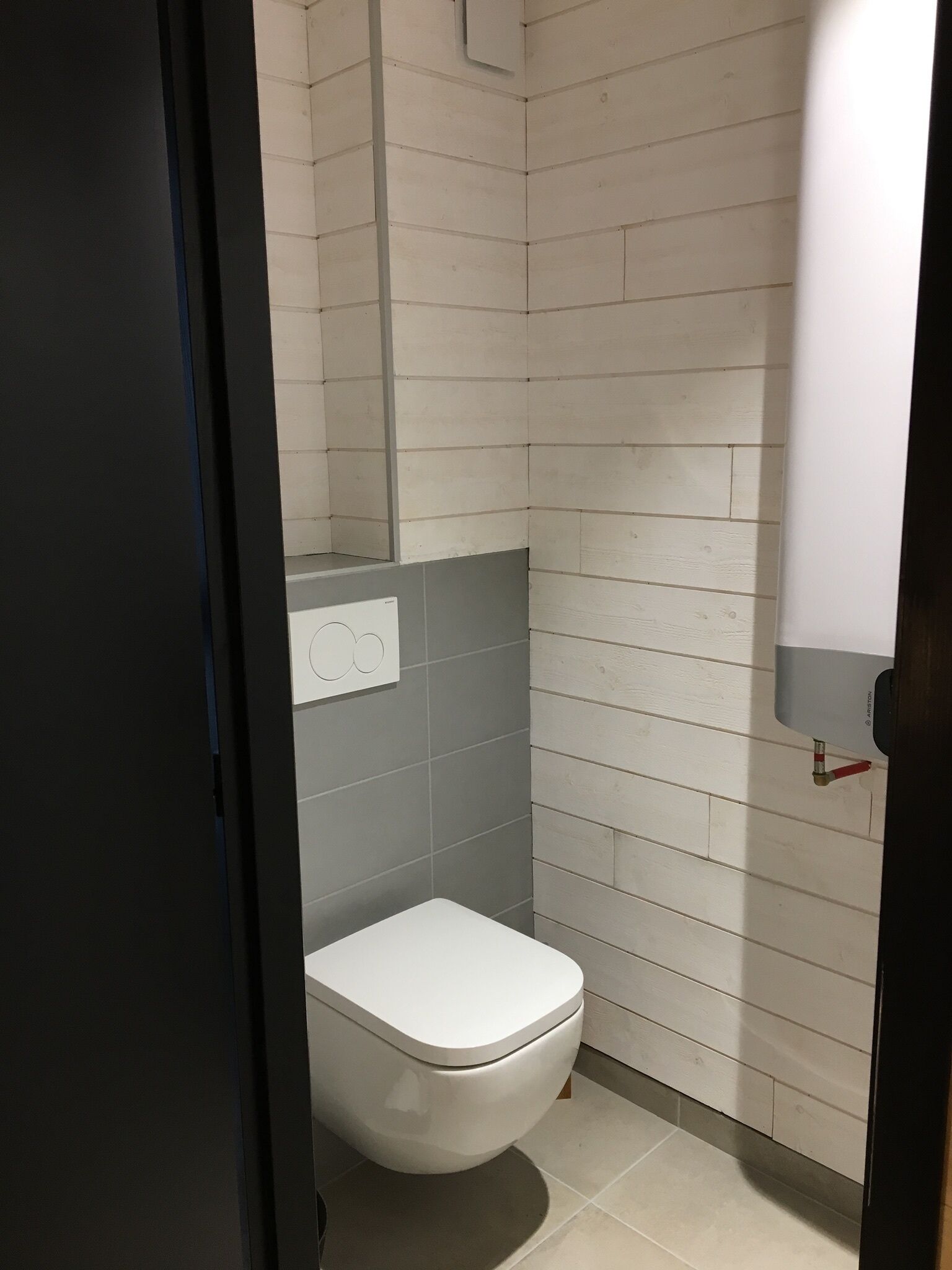 Une petite salle de bain moderne avec des toilettes blanches, des carreaux gris et un chauffe-eau apparent.