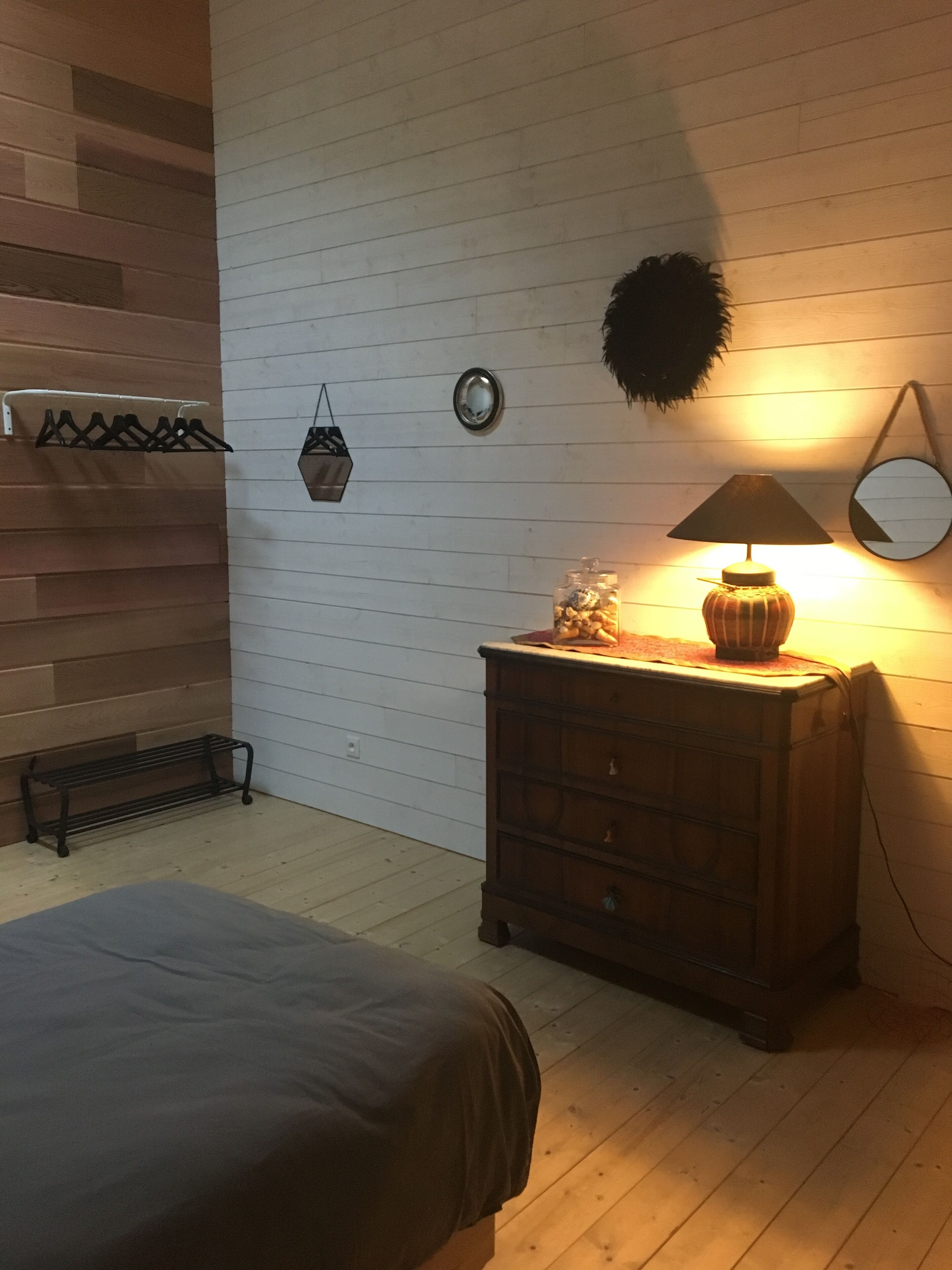 Un dormitorio con suelo de madera, una cama, un aparador, una lámpara, un espejo y una estantería en la pared con ropa.