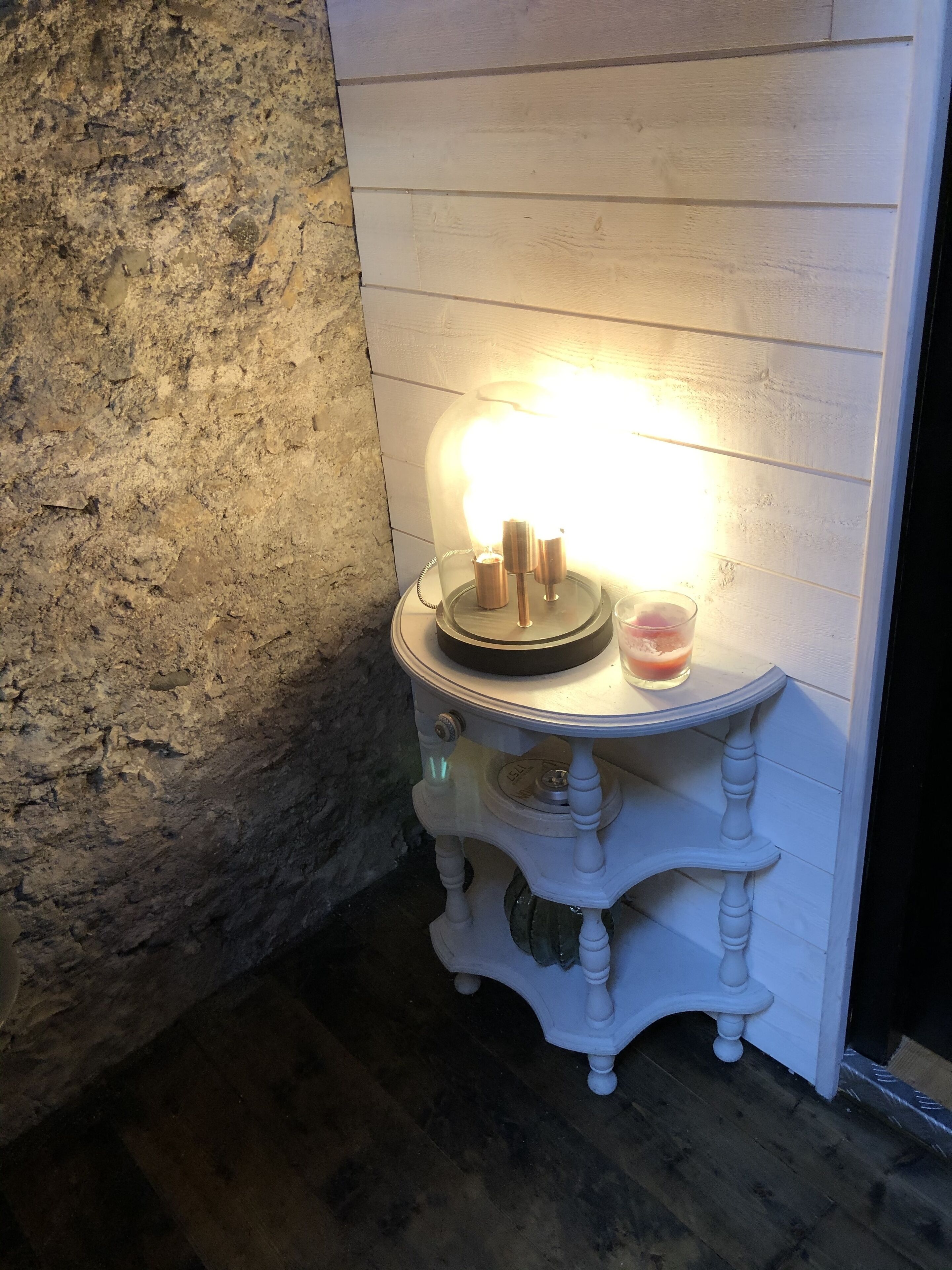 A small, white, antique-style table with a glass top, a candle holder, and a glass container with a pink substance, placed against a textured stone wall and white wooden paneling.