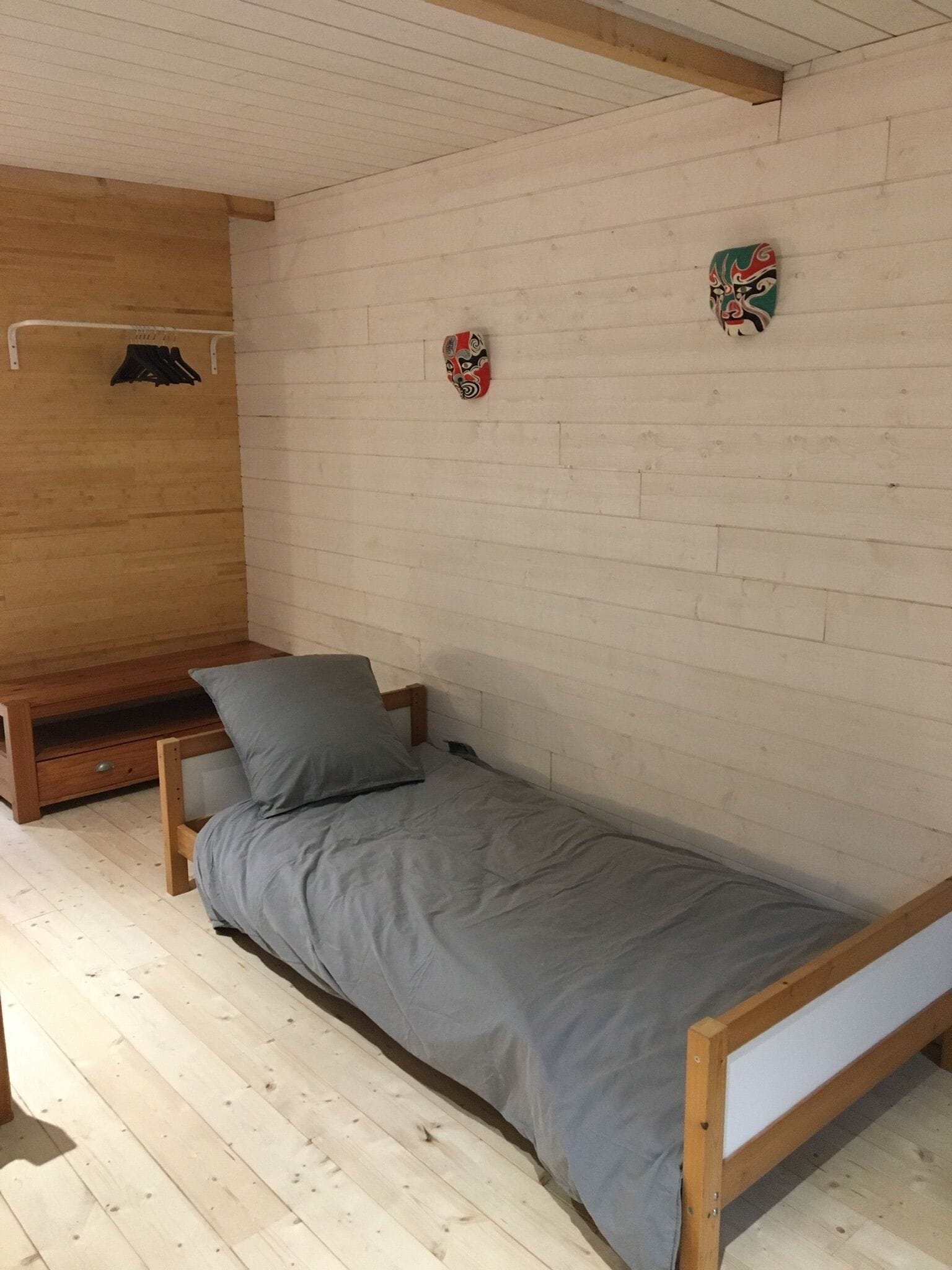 A small, single-bed room with a wooden headboard, grey bedding, and two decorative masks on the wall.