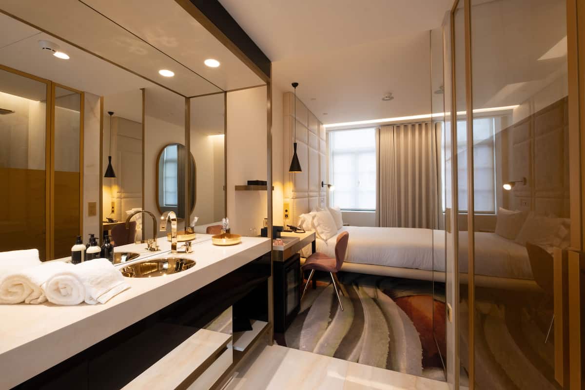 Ein modernes Hotelzimmer mit großem Spiegel, einem Doppelbett, einem Schreibtisch mit Stuhl, einem großen Fenster mit Vorhängen sowie einem Badezimmer mit Waschbecken und Spiegel.