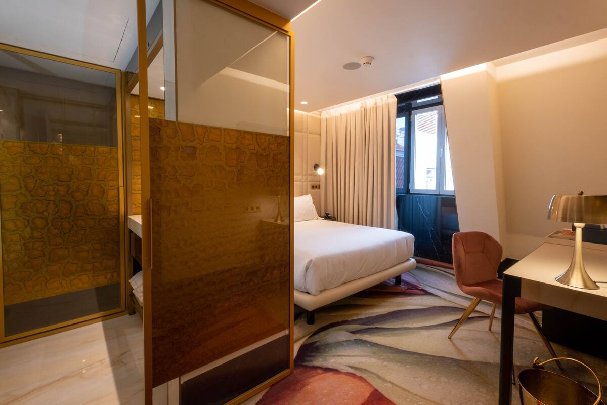 Ein Hotelzimmer mit Bett, Schreibtisch, Stuhl, großem Fenster mit Vorhängen und einem gemusterten Teppich.