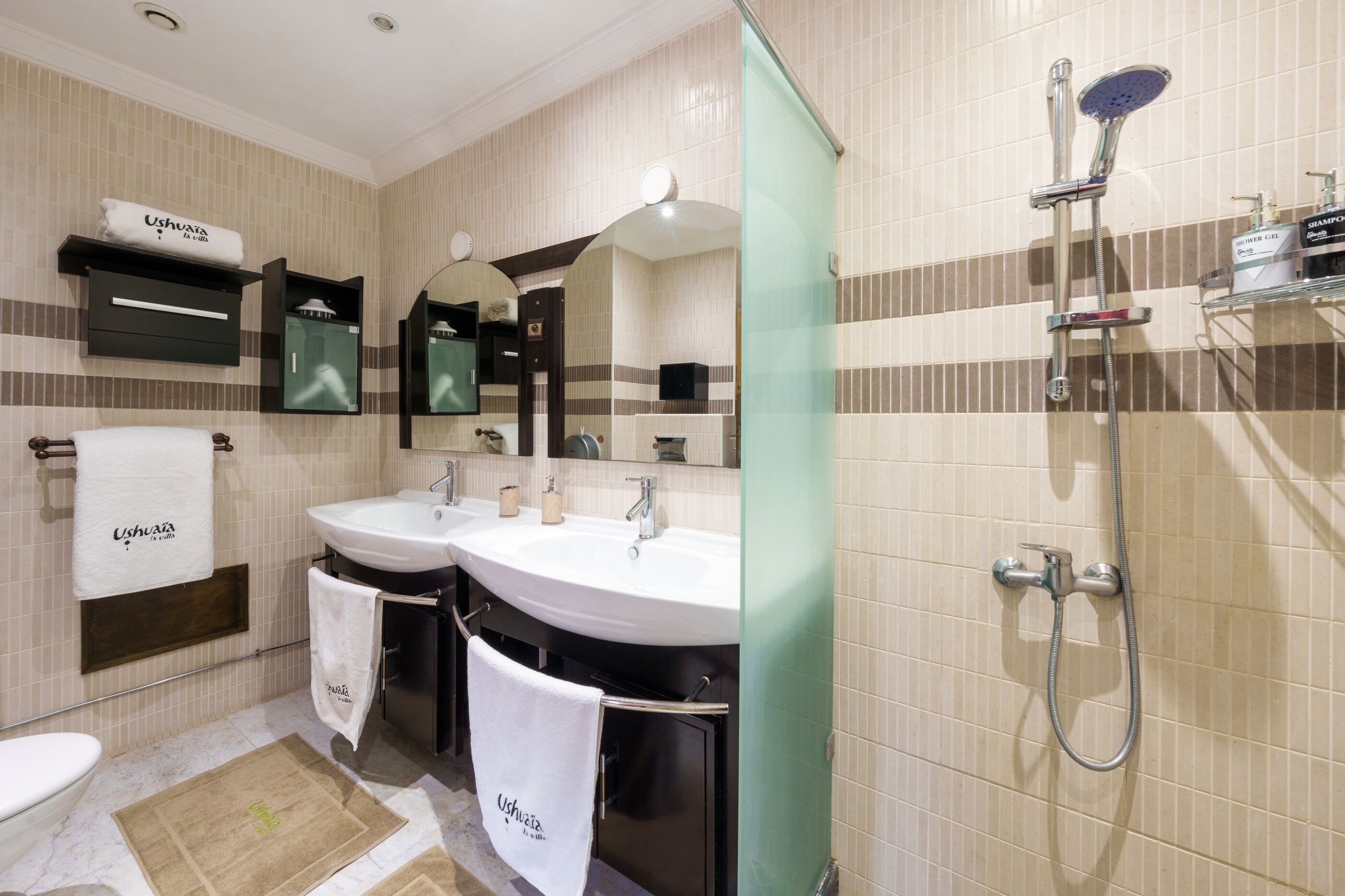 Un baño con dos lavamanos, ducha y espejo. Hay toallas colgadas en la pared y un estante con artículos de aseo.