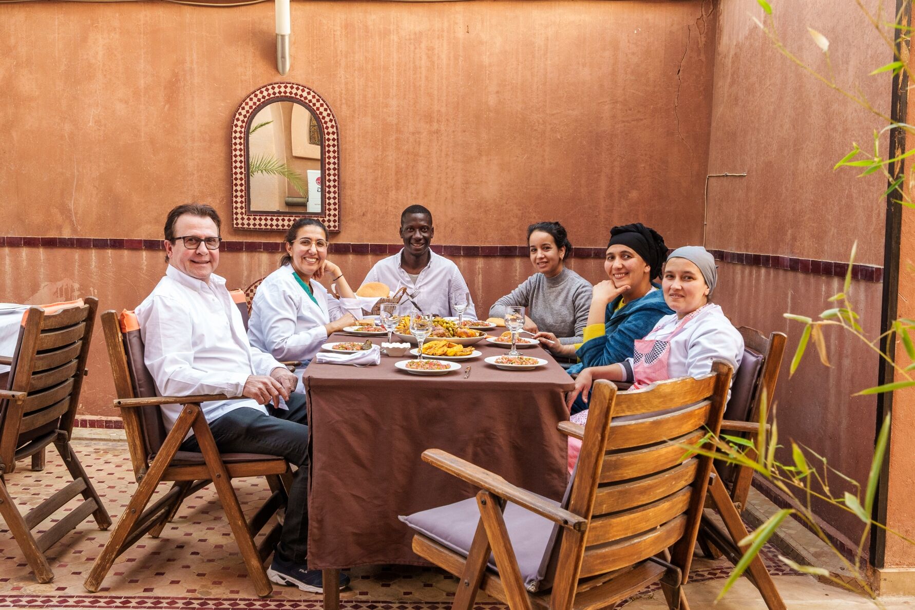 Un grupo de personas sentadas alrededor de una mesa con diferentes platos, en una habitación con sillas de madera y piso de baldosas.