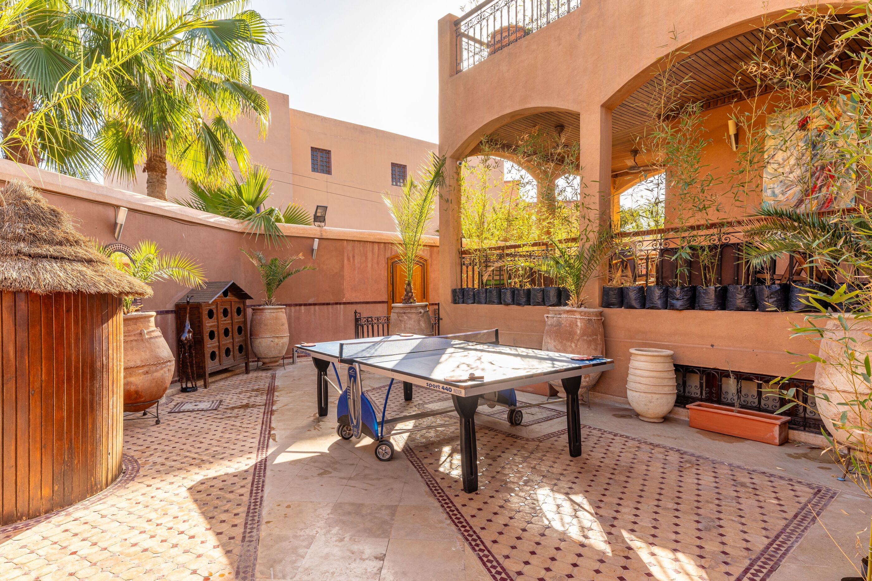 A courtyard with a ping pong table, potted plants, and a thatched-roof structure.