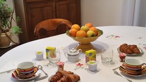 A round dining table with a white tablecloth, a fruit bowl, plates of croissants, and various breakfast items. - Chambres d'hôtes Elisa & Etienne (Manosque)