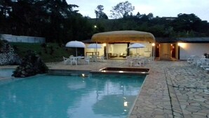 A pool area with a stone patio, white lounge chairs, and umbrellas. - Hotel Itamaracá (Miguel Pereira)