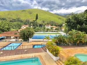 Un complexe hôtelier doté de plusieurs piscines, d’une terrasse en bois et offrant une vue sur une colline où se trouvent des maisons au loin.